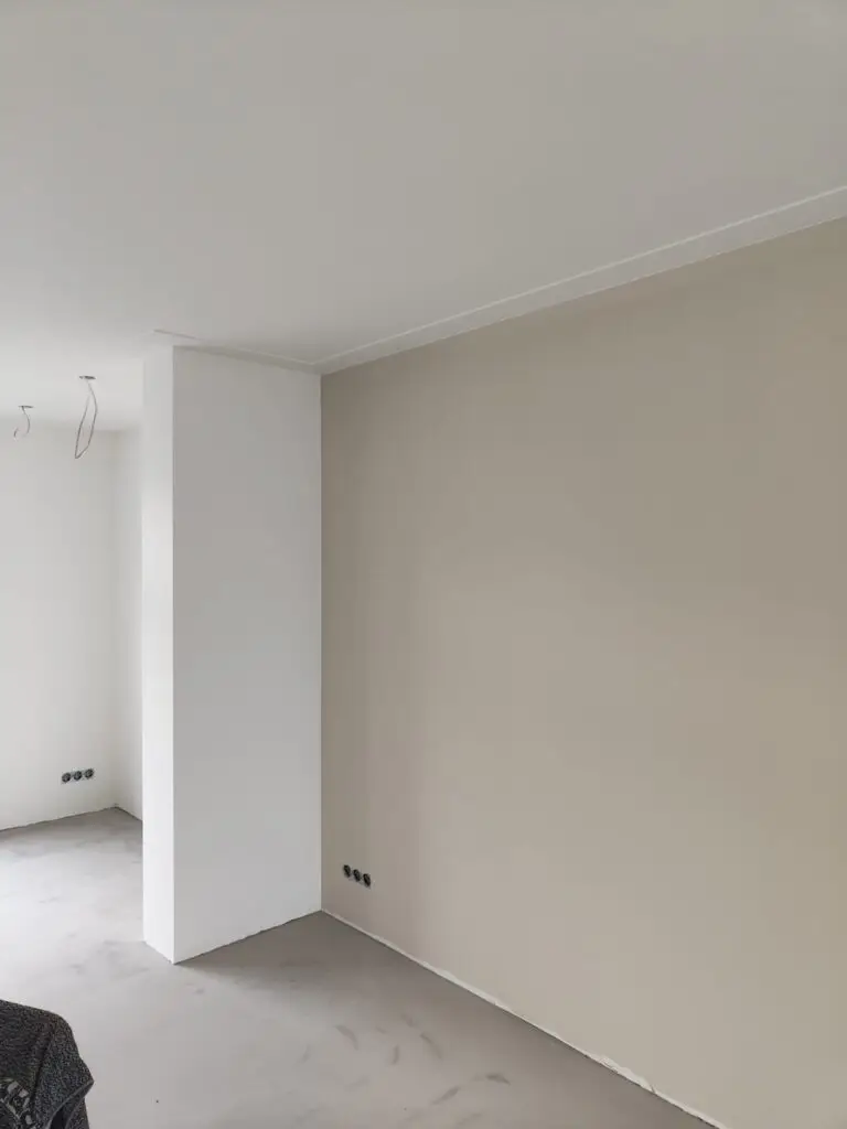 Woonkamer in aanbouw met strakke witte muren, beige accentmuur en onafgewerkte vloeren.